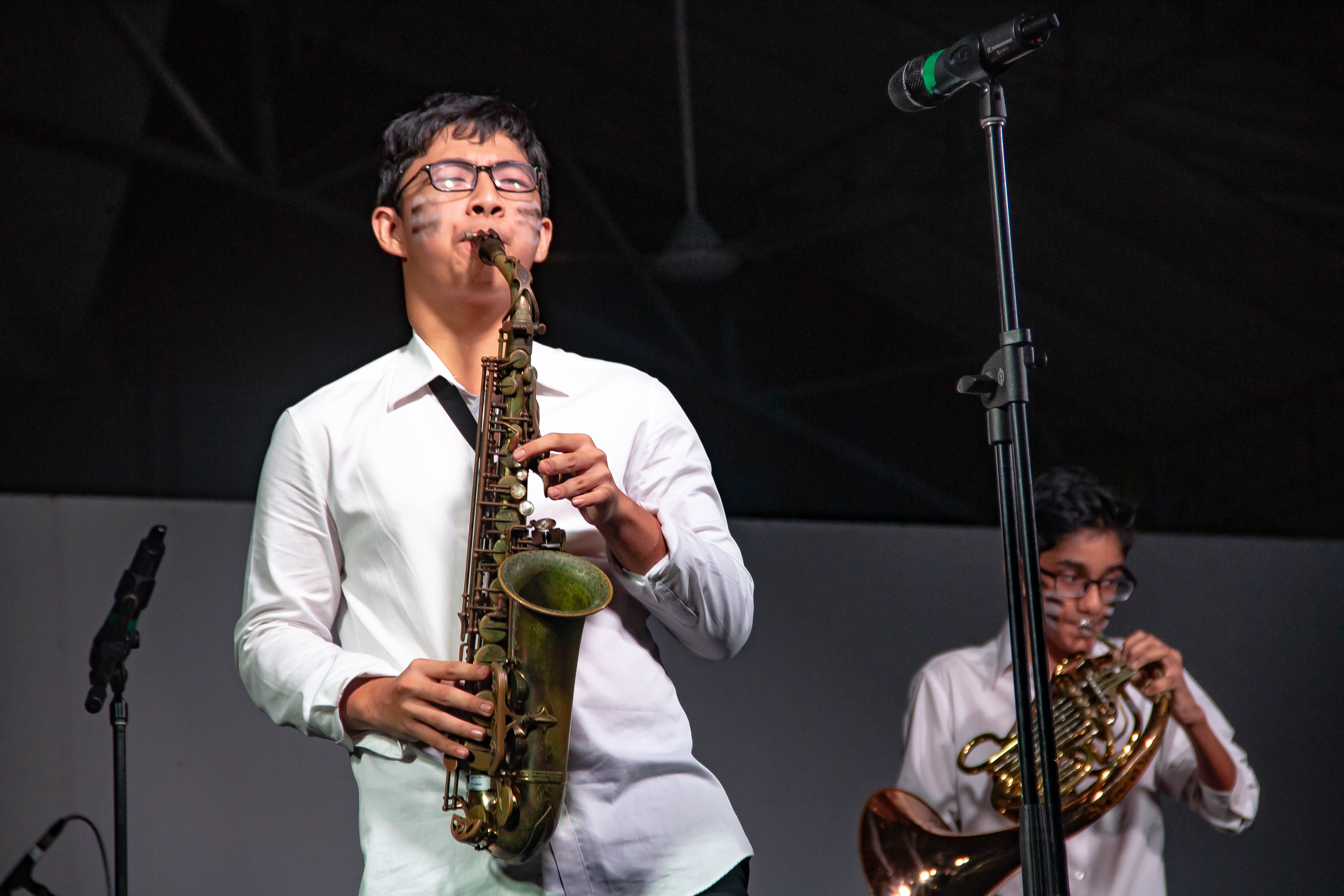 Two young musicians in white shirts perform on stage, one playing saxophone and one playing French horn.